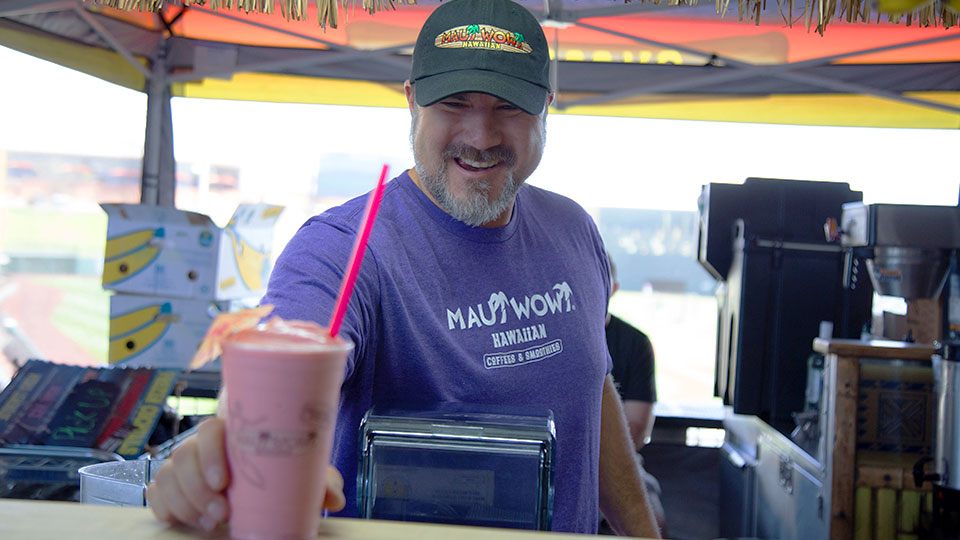 A male franchisee in a purple Maui Wowi t-shirt slides a delicious smoothie across the counter and smiles.