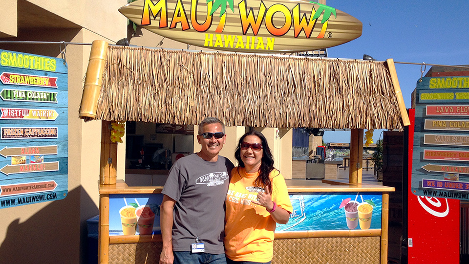 A man and woman, Maui Wowi franchisees, standing in front of a K-Cart, smiling in the sun and giving a cool, cowabunga hand gesture.