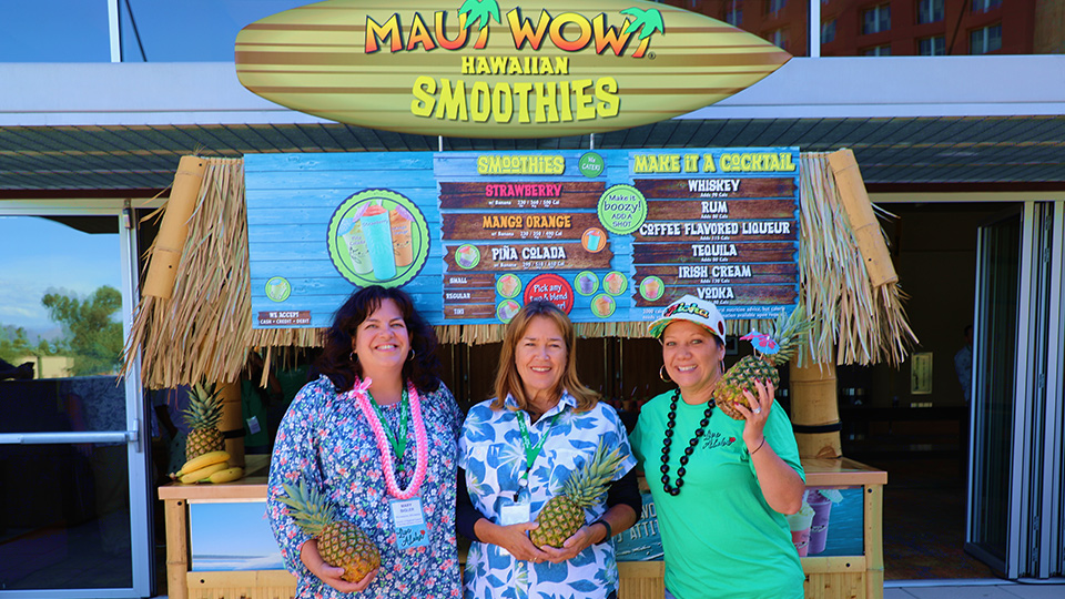 Three women standing in front of a Maui Wowi K-Cart, wearing Hawaiian shirts and holding pineapples.